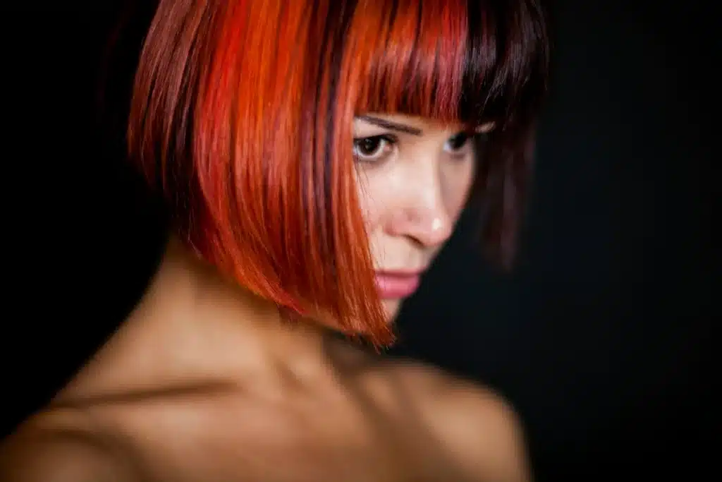 A woman with a glossy copper-red bob and straight fringe against a dark background.