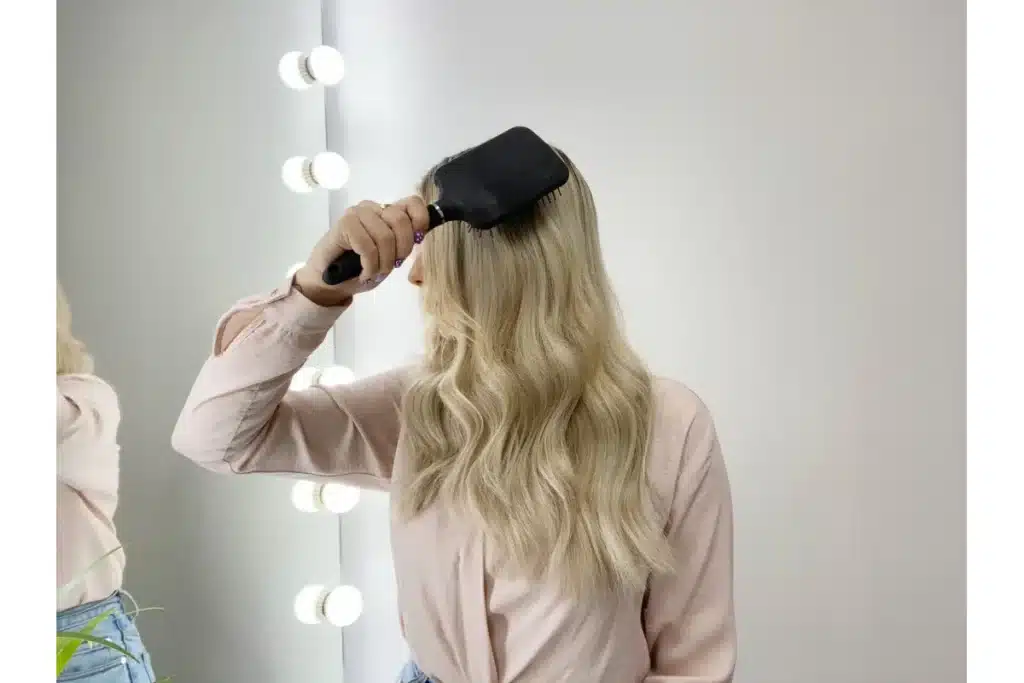 A blonde woman holds a black paddle brush beside long hair.