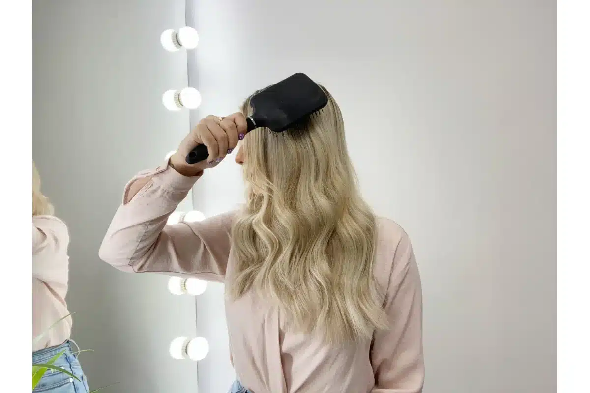A blonde woman holds a black paddle brush beside long hair.