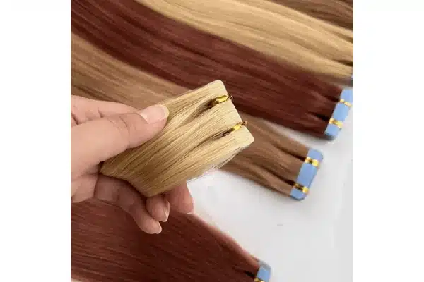 A hand holds blonde tape in extension tabs, with brown and auburn pieces visible behind them.