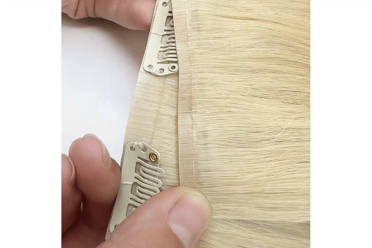 Hands hold a blonde clip-in hair piece to show its narrow seam and attachment clips.